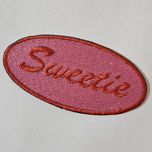 Könnte beinhalten: Ein rosa gestickter Aufnäher mit dem Wort "Sweetie" in Schreibschrift. Der Aufnäher hat einen roten Rand und ist oval.
