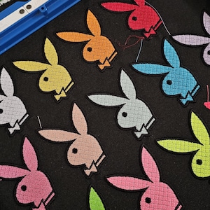 May include: A blue embroidery hoop containing black fabric with multiple embroidered rabbit head logos in various colors including red, orange, yellow, light blue, light gray, pink, lime green, and lavender, each featuring a small bowtie detail.