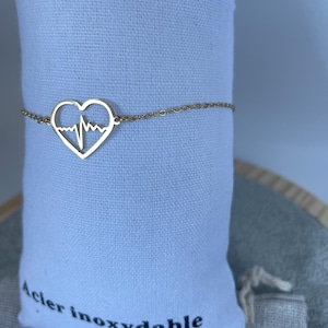 Peut inclure: Un bracelet à chaîne doré doté d'un pendentif central en forme de cœur avec un motif de ligne d'électrocardiogramme intégré. La fine chaîne métallique est fixée de chaque côté du cœur, formant un accessoire de poignet continu.