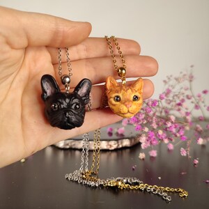 May include: Two animal-themed pendant necklaces. One depicts a black French bulldog, the other an orange tabby cat. Both pendants hang from fine chains. The necklaces are held in a hand against a soft background.