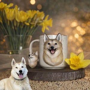 May include: A cream-coloured ceramic mug featuring a 3D dog face, a matching handle, and a small dog figurine. The mug is displayed on a wooden base, with a yellow daffodil and a vase of daffodils in the background. The dog in the image is a husky.