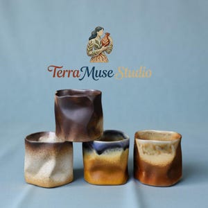 Pode incluir: Quatro canecas de cerâmica artesanais com um design texturizado único. As canecas apresentam uma forma quadrada com laterais dobradas e uma variedade de esmaltes em tons terrosos, incluindo castanho, bege e azul. O logotipo Terra Muse Studio é visível.