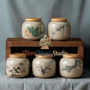 Puede incluir: Cinco tarros de cerámica con tapas de madera se exhiben en una estantería de madera. Los tarros son de color beige con diseños florales y botánicos pintados a mano en verde, rosa y negro. El texto "Terra Muse Studio" es visible.