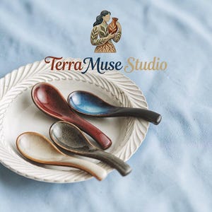 Puede incluir: Cuatro cucharas de cerámica en tonos rojo, azul, gris y crema descansan sobre un plato blanco texturizado. Las cucharas tienen un acabado liso y brillante. La imagen incluye el texto "Terra Muse Studio" y un logotipo.