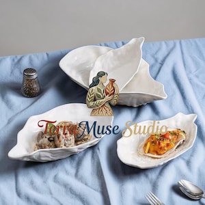 May include: White, leaf-shaped ceramic serving dishes. One dish holds seafood, another has a cooked dish, and a third is empty. A decorative wooden figure of a woman holding a vase is in the center. The text "Terra Muse Studio" is on one dish.