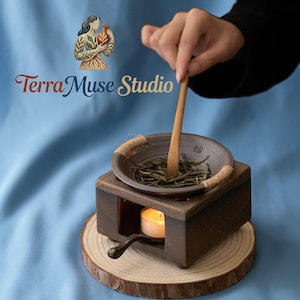 May include: A dark brown ceramic tea warmer with a small candle inside, a ceramic bowl with tea leaves, and a wooden spoon. The warmer sits on a wooden slice. The text "Terra Muse Studio" is in the upper left corner.