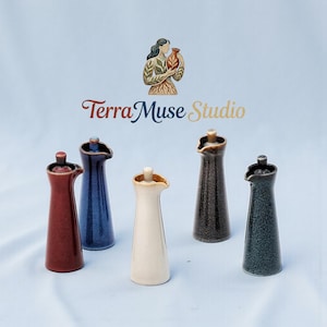Puede incluir: Una colección de dispensadores de aceite de cerámica en varios colores, incluyendo rojo, azul, crema, marrón y verde azulado oscuro. Cada dispensador tiene una forma delgada y alargada con un pico y un pequeño tapón. Se ve el logo de Terra Muse Studio.