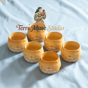 Puede incluir: Seis pequeñas macetas redondas de cerámica de color beige claro con caras de gato de dibujos animados. Están dispuestas sobre una superficie azul claro. El texto "Terra Muse Studio" es visible.