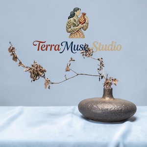 Puede incluir: Un jarrón texturizado de color bronce, con una base ancha y redondeada y un cuello estrecho, que contiene ramas secas con pequeñas hojas marrones. El jarrón está sobre una superficie azul claro. El logo "Terra Muse Studio" es visible.