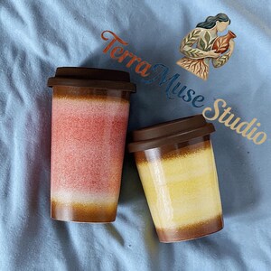 May include: Two ceramic travel mugs with brown lids. One mug is red and white, the other is yellow and brown. The mugs are on a blue surface. The text "Terra Muse Studio" is in the image.