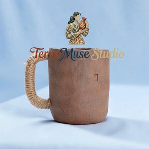 Könnte beinhalten: Eine braune Keramiktasse mit einem Seilgriff. Die Tasse hat eine quadratische Vertiefung an der Seite. Der Text "Terra Muse Studio" ist auf die Tasse geschrieben. Über der Tasse befindet sich eine Illustration einer Frau, die eine Vase hält.