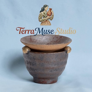 May include: A ceramic bowl set with a textured, dark brown exterior and a lighter brown interior. The set includes a bowl and a lid, with a small golden sphere on the side. The logo "Terra Muse Studio" is displayed above the bowl.