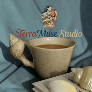 Puede incluir: Una taza de cerámica moteada con un asa en forma de concha, llena de café. La taza está sobre un libro junto a conchas marinas. El texto "Terra Muse Studio" está encima de la taza, con una ilustración de una mujer sosteniendo un jarrón.