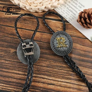 May include: Two bolo ties with black braided cords. One has a silver clasp with "Sophie". The other has a silver and gold oval pendant with the letter "R". They are displayed on a wooden surface.