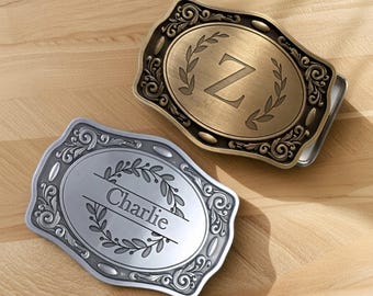 May include: Two decorative metal belt buckles. One is silver with the name "Charlie" engraved, and the other is bronze with the letter "Z". Both have an oval center with a laurel wreath design and ornate borders. They rest on a wooden surface.