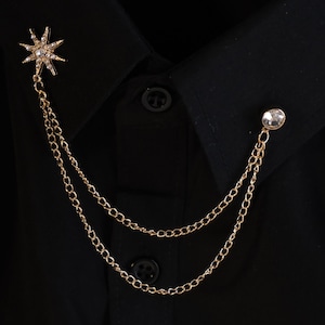 May include: Gold-toned starburst and crystal chain collar pin. The starburst is encrusted with small crystals. The chain drapes down to a crystal-studded button-like accent. The pin is attached to a black shirt.
