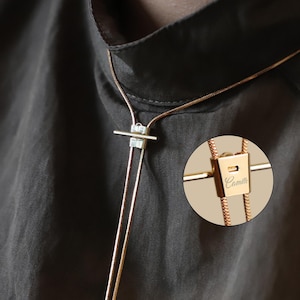 May include: Gold-toned necklace with a rectangular pendant and the name "Camille" engraved. The necklace features a bar and two delicate chains. The background is a dark, solid color.