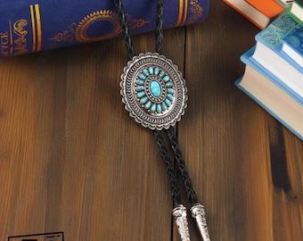 May include: A silver bolo tie with a turquoise stone center and accents. The bolo tie has a braided black leather cord with decorative silver tips. The bolo tie is on a wooden surface with books in the background.