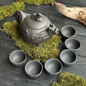 Może przedstawiać: Ciemnoszary ceramiczny zestaw do herbaty z detalami smoka i małą figurką zwierzęcia na pokrywce, otoczony sześcioma pasującymi filiżankami. Zestaw jest prezentowany na zielonym mchu.
