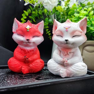May include: Two small, ceramic fox figurines in a seated meditation pose. The fox on the left is red, and the fox on the right is white. Both foxes have closed eyes and a small, pink flower on their foreheads.