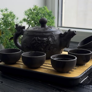 Op de afbeelding: Een zwarte theepot met ingewikkelde drakenontwerpen en vier bijpassende theekopjes op een houten dienblad. De theepot en de kopjes zijn gemaakt van keramiek en hebben een matte afwerking.