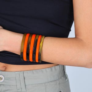 Peut inclure: Une série de six bracelets colorés sur un poignet. Les bracelets alternent entre des teintes orange, noires et marron. Les bijoux sont portés avec un haut tube noir et un pantalon vert clair.