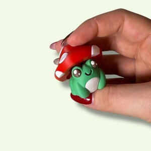 May include: A miniature green frog figure with a white belly and large eyes, topped with a red mushroom cap with white spots. The frog is a whimsical, miniature sculpture.