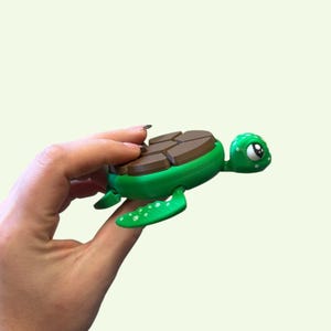 May include: A green and brown turtle-shaped toy with a cartoon design. The turtle has a green body, brown shell, and white spots on its flippers. The turtle's head has large, expressive eyes.