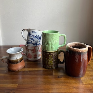 May include: A collection of ceramic mugs in various styles and colors. The mugs include a blue and white landscape design, a pink floral pattern, a green bamboo design, a brown mug with a relief design, and a large brown mug with a textured rim.