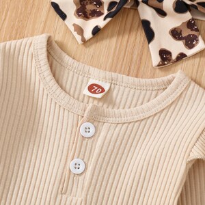 May include: Close-up of a beige ribbed knit baby outfit with two white buttons and a small tag with the number 70. A matching headband with a brown and black animal print is visible in the background. The outfit is made of a soft, ribbed fabric.