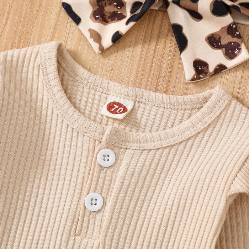 May include: Close-up of a beige ribbed knit baby outfit with two white buttons and a small tag with the number 70. A matching headband with a brown and black animal print is visible in the background. The outfit is made of a soft, ribbed fabric.