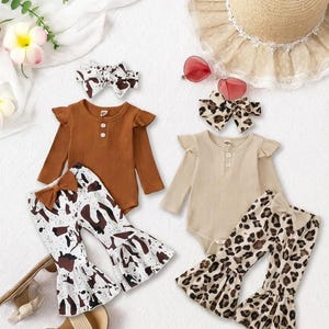 Könnte beinhalten: Zwei Baby-Outfits mit passenden Stirnbändern und Schlaghosen. Ein Set ist braun mit Kuhdruck, das andere beige mit Leopardenmuster. Ein Strohhut und herzförmige Sonnenbrillen sind ebenfalls im Bild.