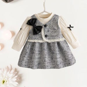 May include: A baby girl's dress with a white top and a gray tweed skirt and vest. The dress features a large black bow, black buttons, and small black bows on the sleeves. The dress is hanging on a white hanger.