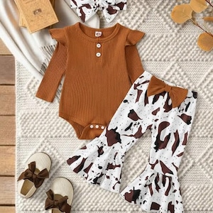 May include: A baby clothing set featuring a rust-colored bodysuit with ruffle sleeves and a white and brown cow print pants. The set includes a matching headband and shoes with brown bows. The outfit is displayed on a textured surface.