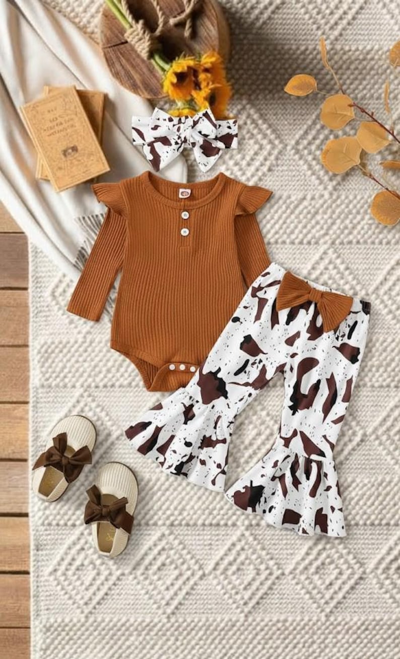 May include: A baby clothing set featuring a rust-colored bodysuit with ruffle sleeves and a white and brown cow print pants. The set includes a matching headband and shoes with brown bows. The outfit is displayed on a textured surface.