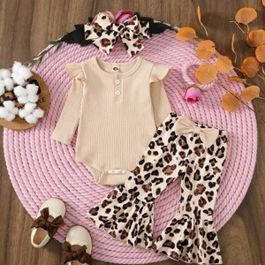 May include: A beige ribbed bodysuit with ruffle sleeves and three buttons, paired with leopard print bell-bottom pants and a matching bow headband. The outfit includes beige bow-adorned shoes. The ensemble is designed for infants.