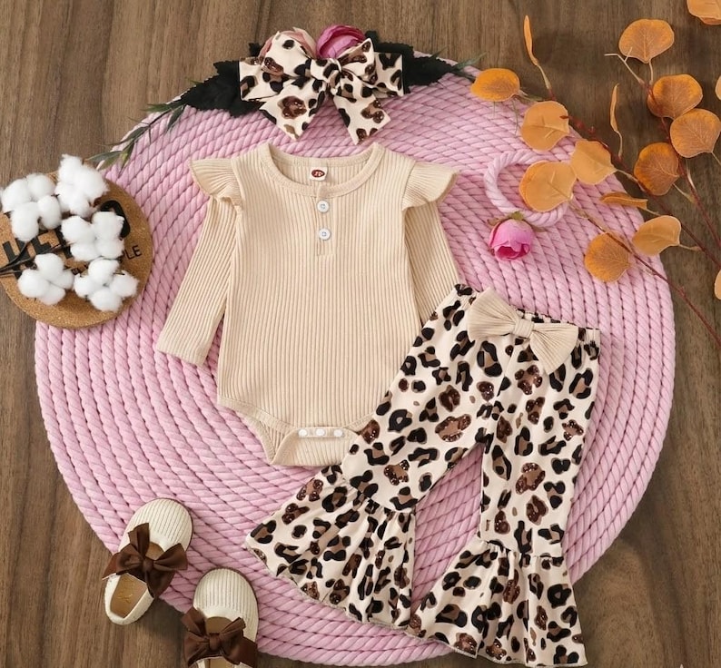 May include: A beige ribbed bodysuit with ruffle sleeves and three buttons, paired with leopard print bell-bottom pants and a matching bow headband. The outfit includes beige bow-adorned shoes. The ensemble is designed for infants.