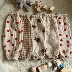 May include: Five pairs of baby pants in various patterns, including red hearts, brown plaid, brown teddy bears, small white flowers, and red ladybugs. The pants are made of a soft, textured fabric and have elasticated cuffs and waistbands.
