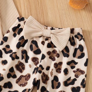 May include: Close-up of a beige and brown leopard print pants with a matching beige bow. The pants are made of a soft material and are designed for comfort. The bow is a prominent feature, adding a touch of style.