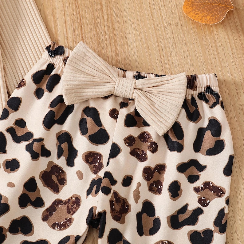 May include: Close-up of a beige and brown leopard print pants with a matching beige bow. The pants are made of a soft material and are designed for comfort. The bow is a prominent feature, adding a touch of style.