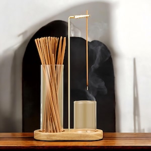 May include: A wooden incense holder with two glass cylinders. One cylinder holds a bundle of incense sticks, and the other holds a single incense stick that is burning. The holder has a gold metal stand with a hook for hanging the incense stick.