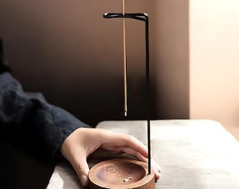 Könnte beinhalten: Ein schwarzer Metall-Räucherstäbchenhalter mit einem Holzsockel. Der Halter hat eine einzelne, dünne Metallstange, die nach oben ragt und für das Halten eines Räucherstäbchens ausgelegt ist. Das Räucherstäbchen hängt an der Stange.