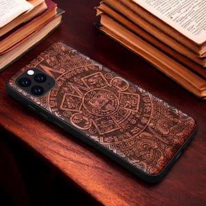 May include: A brown wooden phone case with an intricate carved design of a Mayan calendar. The case has a black border and a camera cutout.