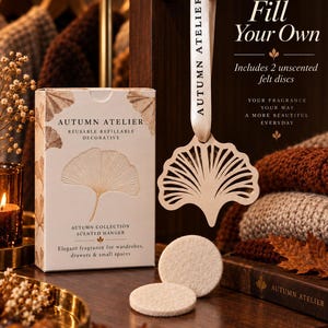 May include: A white rectangular box labeled Autumn Atelier Reusable Refillable Decorative Autumn Collection Scented Hanger, a ginkgo leaf-shaped wooden hanger on a branded ribbon, and two round beige felt discs for fragrance application.