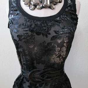 May include: A black sheer top with a floral pattern and a black leather belt. The top has a large silver statement necklace with large, faceted stones.