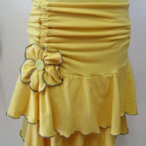 May include: A yellow skirt with a ruffled hem and a large flower applique. The skirt has a gathered waistband and is made of a soft, lightweight fabric.