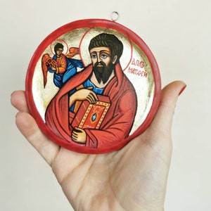 Puede incluir: Icono religioso redondo pintado a mano que representa a un santo con una túnica roja sosteniendo un libro. El icono tiene un fondo dorado y un borde rojo. Un ángel está representado en el fondo. Se adjunta un pequeño lazo de metal para colgar.