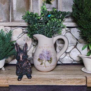 May include: A ceramic pitcher with a floral design and the text "The Botanical Society" and "Hydrangea flower". A small bronze rabbit figurine sits in front of the pitcher.