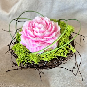 Op de afbeelding: Een roze en witte pioenroos ligt in een klein, geweven bruin nest gevuld met felgroen mos. Groene stelen buigen zich om de bloem en creëren een natuurlijke, decoratieve opstelling.