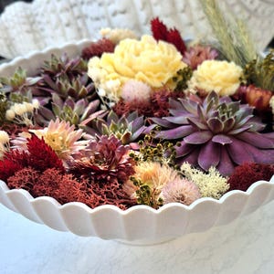 Peut inclure: Un bol blanc à bord festonné rempli d'un assortiment de fleurs séchées et de plantes succulentes. La composition comprend des succulentes violet foncé, des bougies en forme de fleurs crème, de la mousse rouge et des fleurs séchées roses et blanches.
